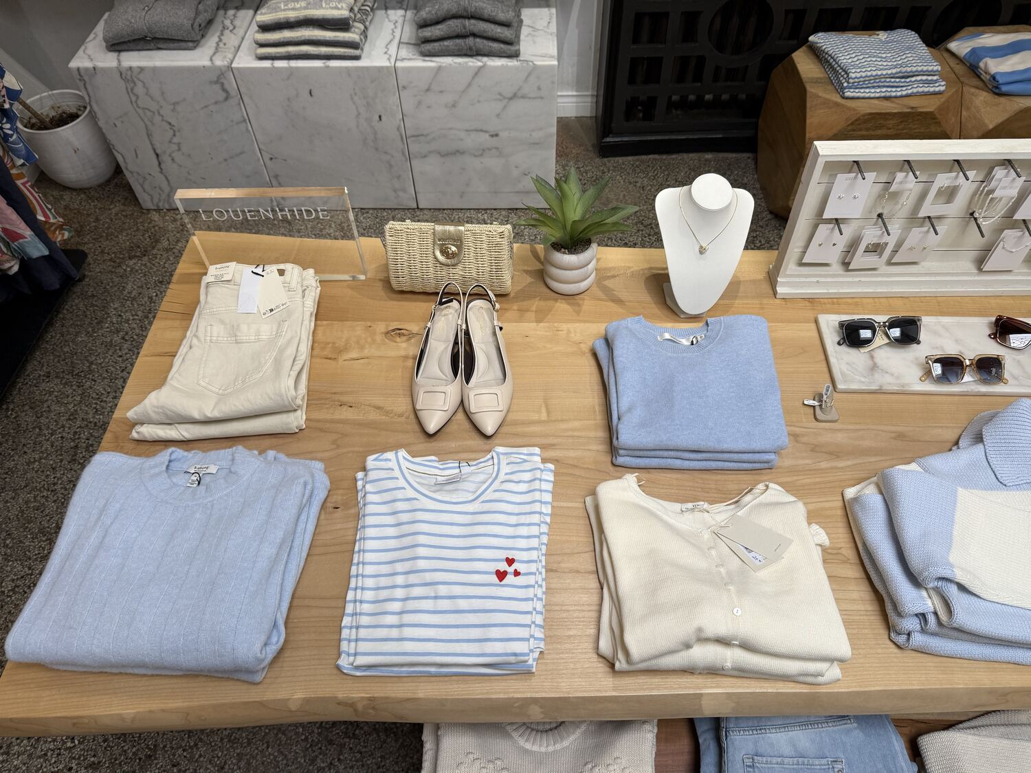 A wooden table styled in powder blue and ivory, with folded knits, slingback shoes, a woven bag, a jewellery bust and sunglasses